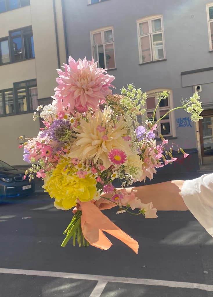 A loose pastel summer bouquet with pink and cream dahlias, daisies and yellow peonies, held up in the street