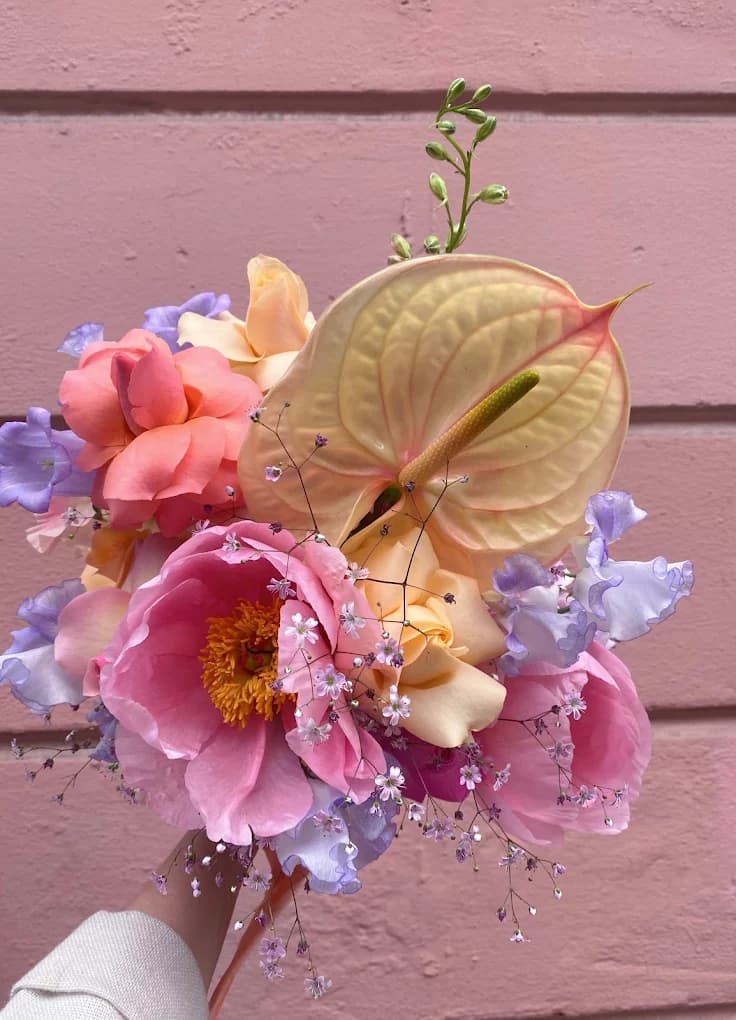 A small bouquet of pink peonies, coral roses and lilac sweet peas held against a pink brick wall